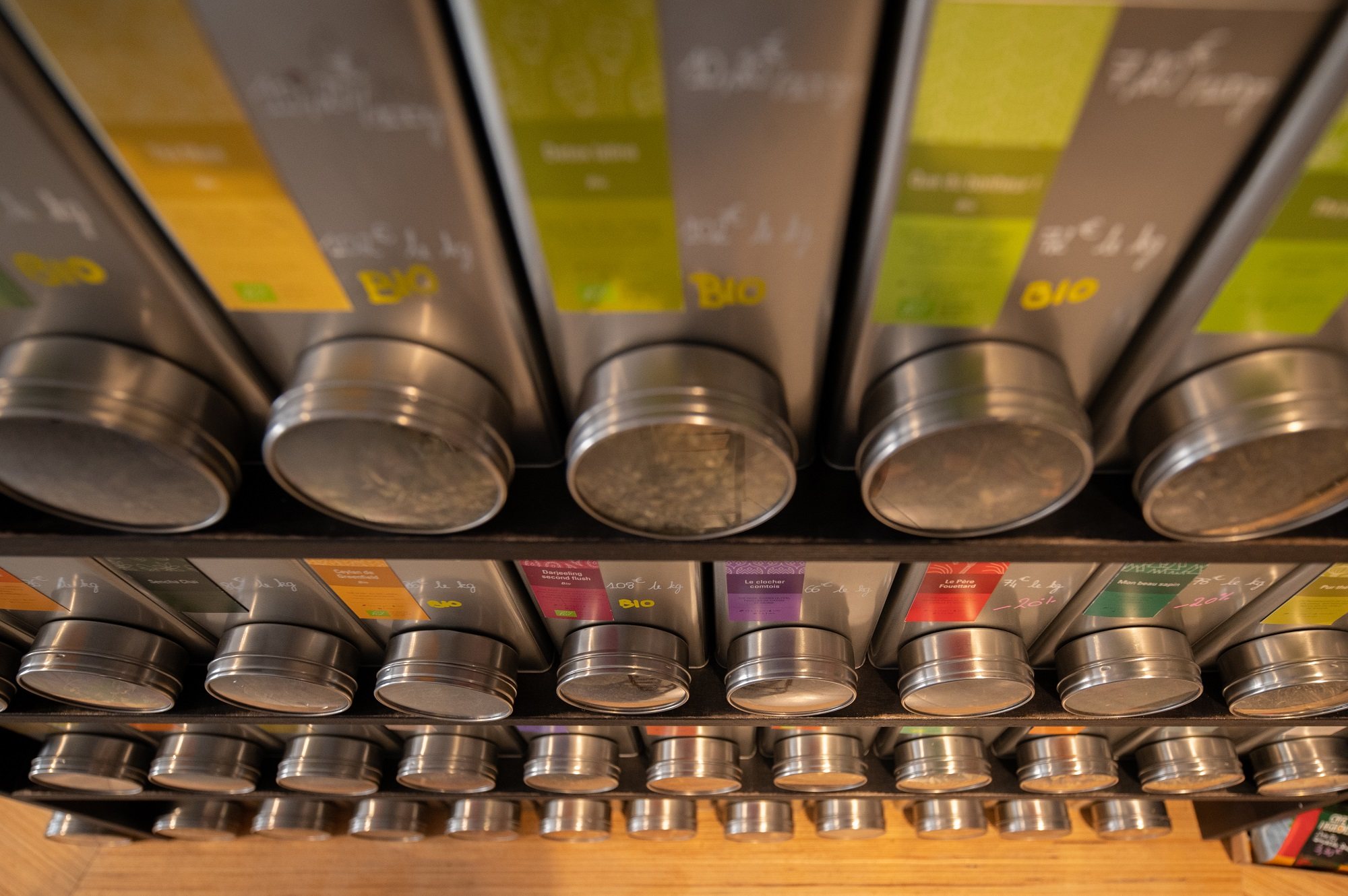 Vue en contre-plongée d'étagères garnies de boîtes métalliques cylindriques à étiquettes colorées, plusieurs portant la mention BIO, pour la vente de thé en vrac.