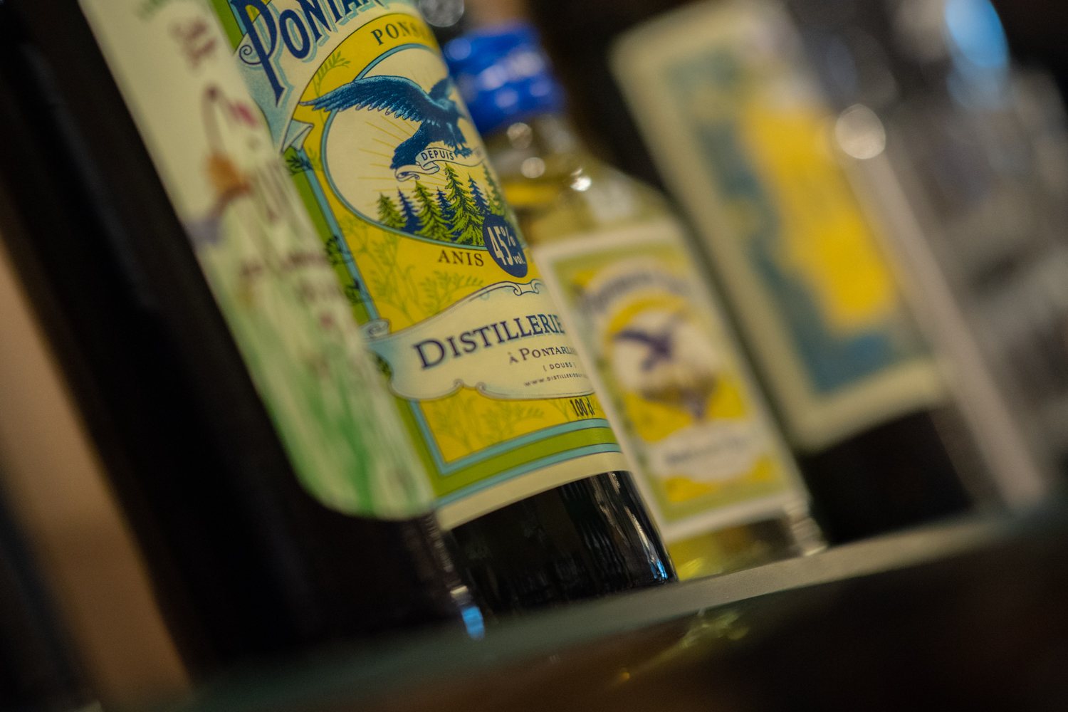 Gros plan en perspective de bouteilles d'anis à l'étiquette jaune et bleue ornée d'un aigle, posées sur une étagère en verre.
