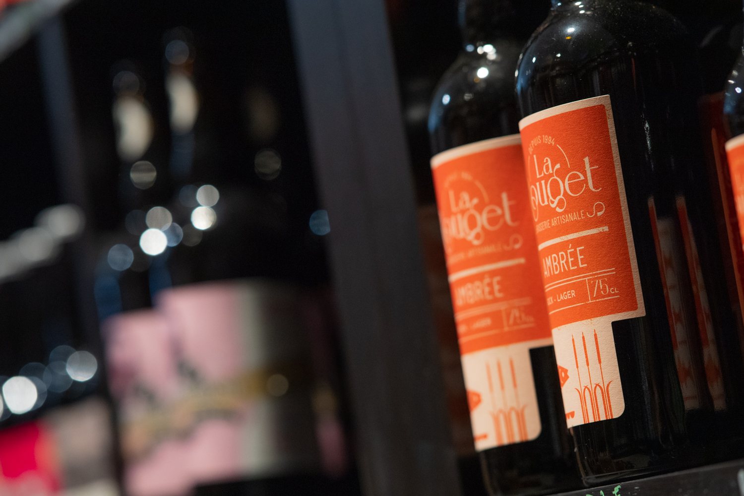 Gros plan sur des bouteilles de bière ambrée de 75 cl à étiquette orange, alignées sur une étagère sombre avec un arrière-plan flou.