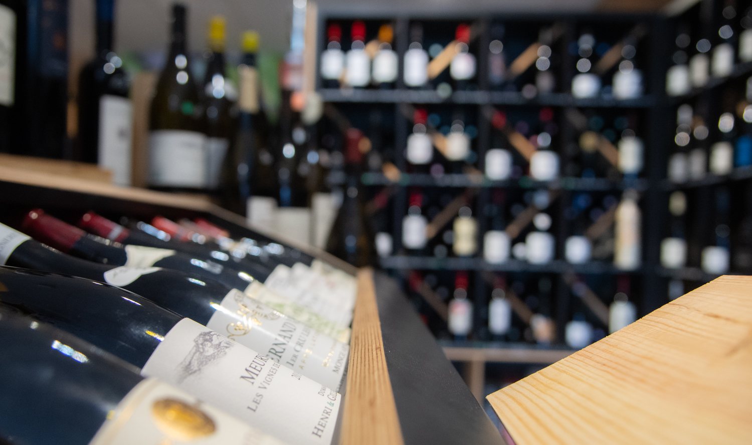 Des bouteilles de vin rouge sont inclinées sur une étagère en bois au premier plan, devant un grand casier rempli de bouteilles floues.