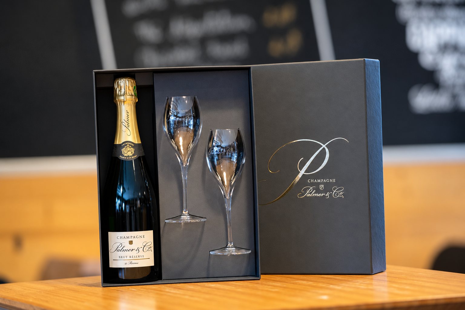 Un coffret noir ouvert contenant une bouteille de champagne Palmer & Co Brut Réserve et deux flûtes en cristal, posé sur une table en bois.
