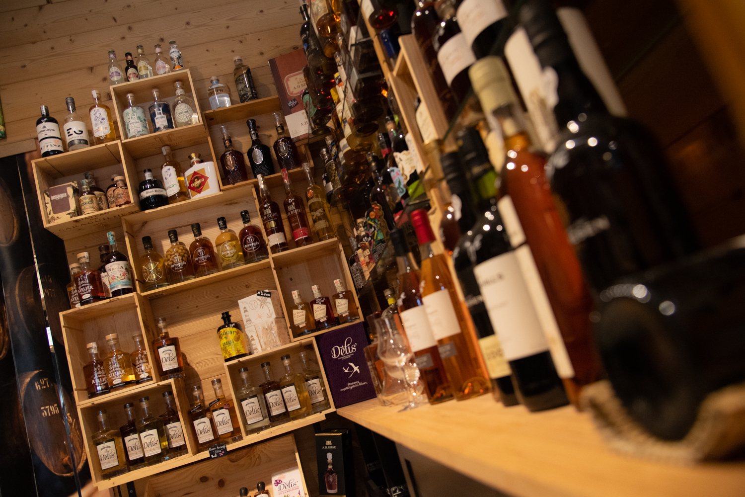 Cave à spiritueux avec des casiers en bois remplis de bouteilles de rhum, whisky et liqueurs, et des bouteilles de vin alignées sur un comptoir au premier plan.