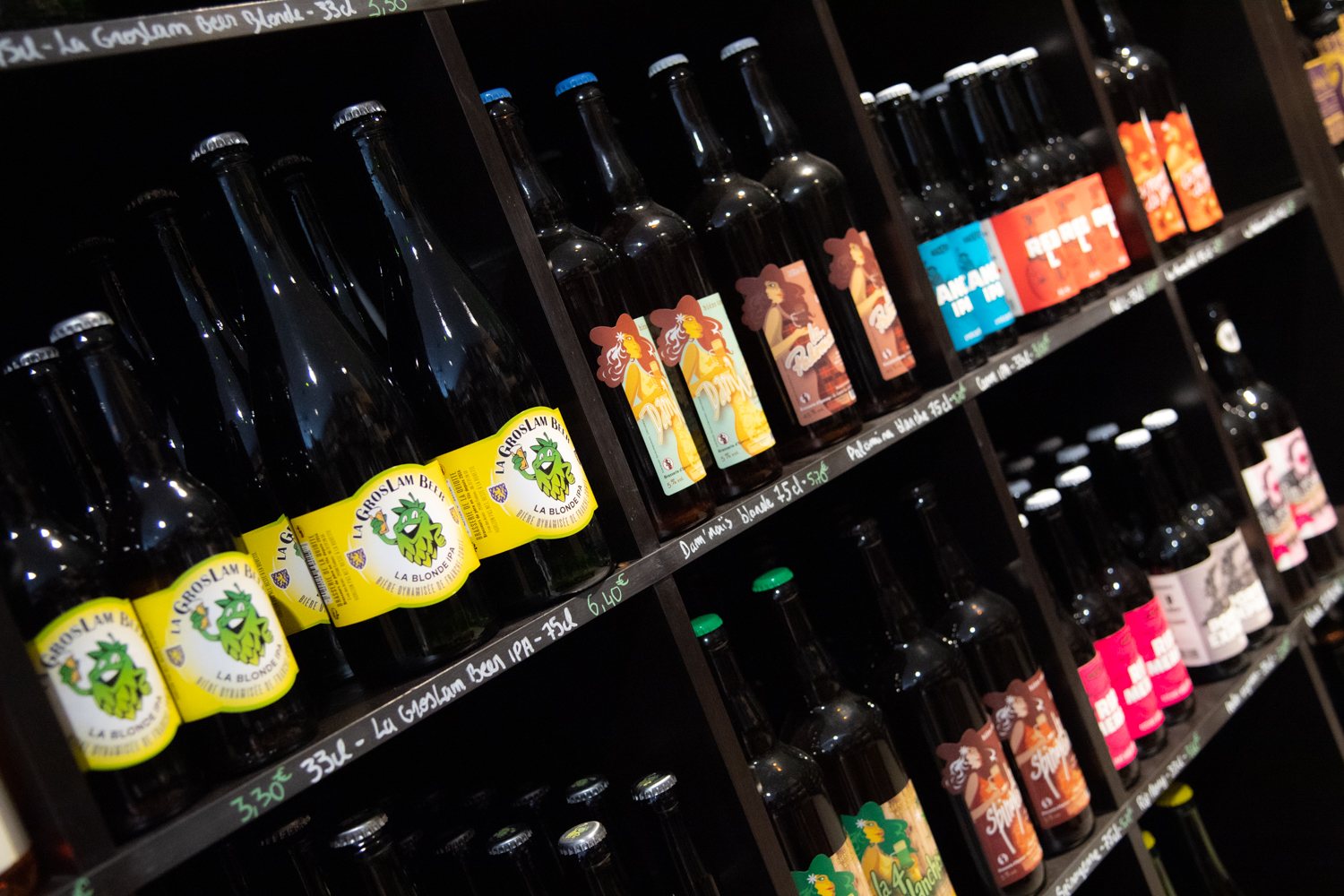 Des étagères noires inclinées présentent de nombreuses bouteilles de bières artisanales aux étiquettes jaunes, oranges, bleues et roses, avec des prix écrits à la craie.