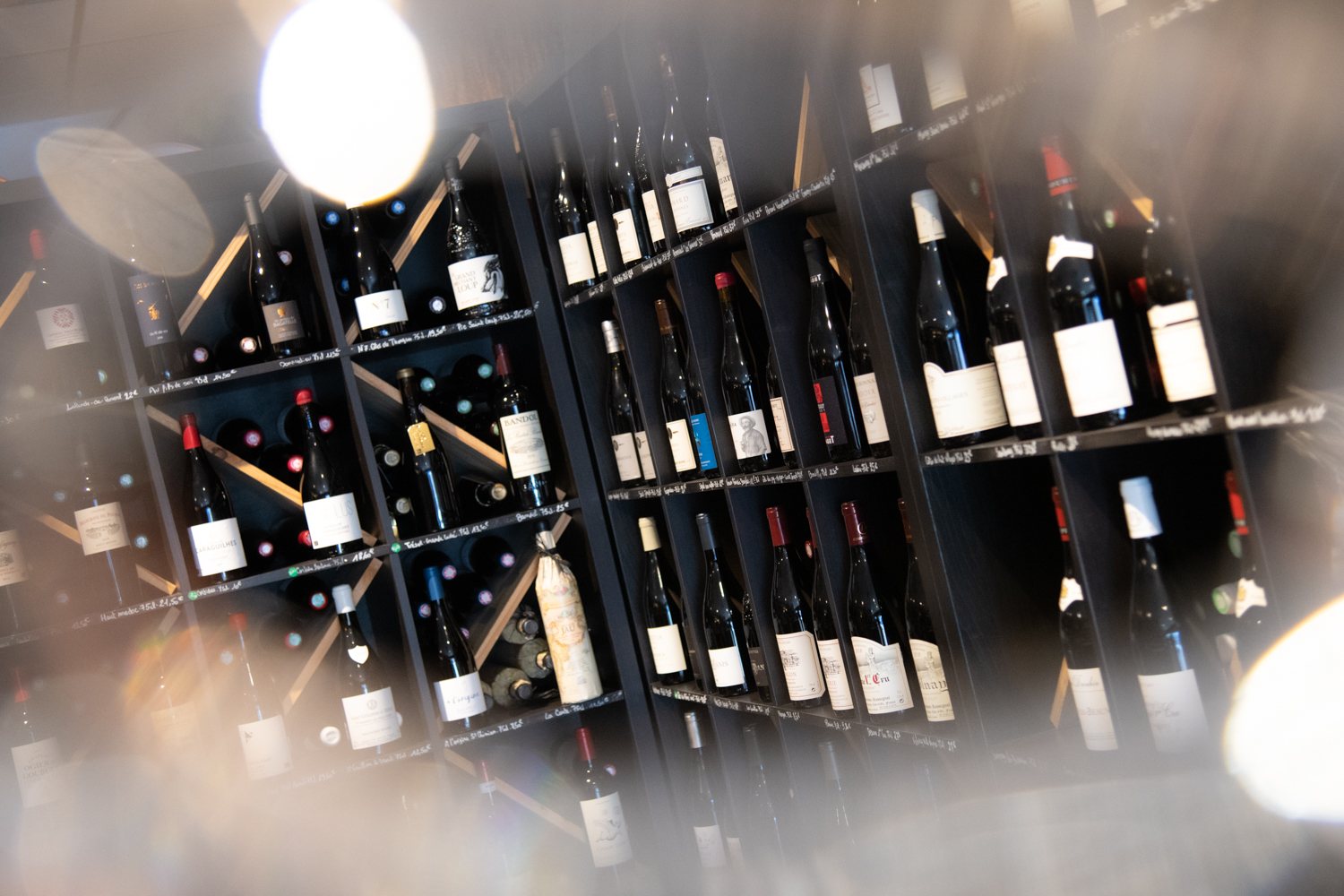 Des bouteilles de vin sont rangées sur des étagères en bois sombre, avec des inscriptions à la craie sur les tablettes et des reflets de lumière floue au premier plan.