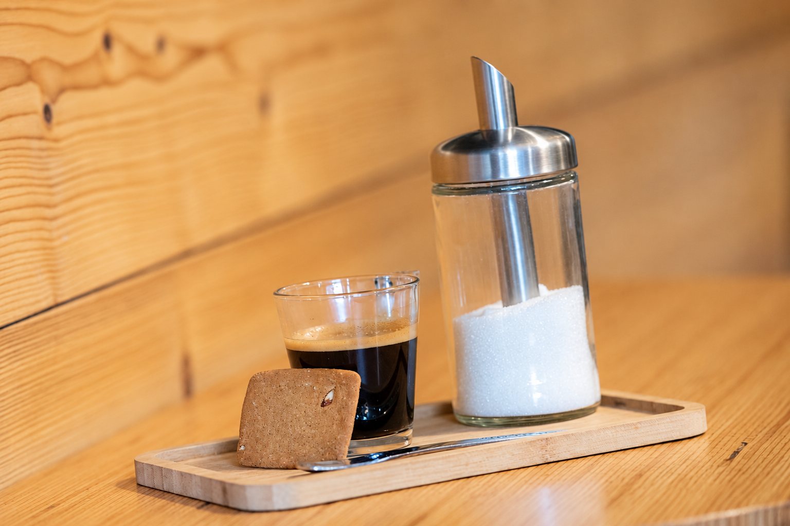 Un petit verre d'expresso accompagné d'un biscuit, d'une cuillère et d'un sucrier verseur en verre, posés sur un plateau en bambou devant un mur en bois clair.