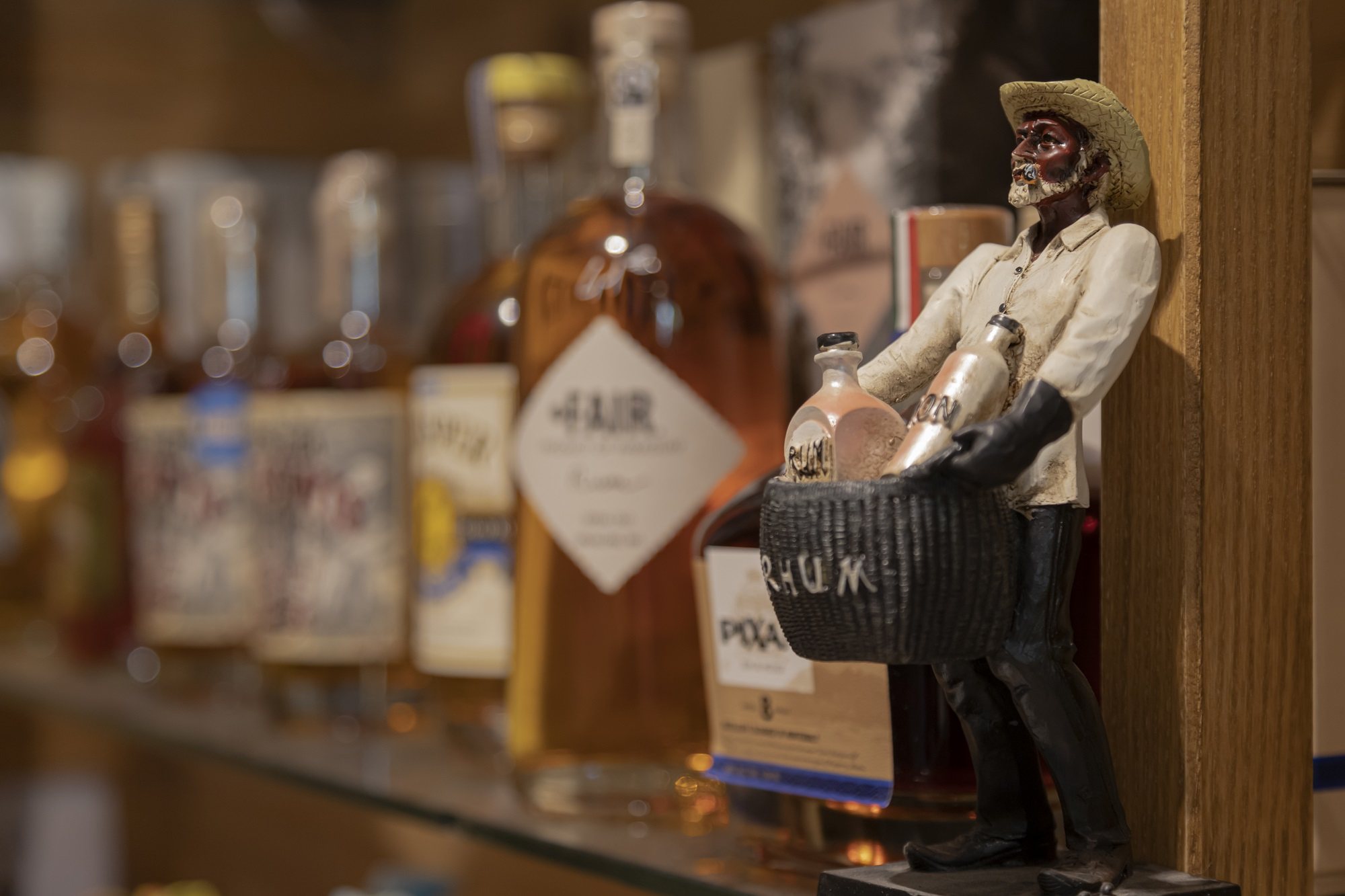Une figurine d'homme coiffé d'un chapeau de paille portant un panier marqué RHUM se tient près d'une étagère de bouteilles de spiritueux floues.