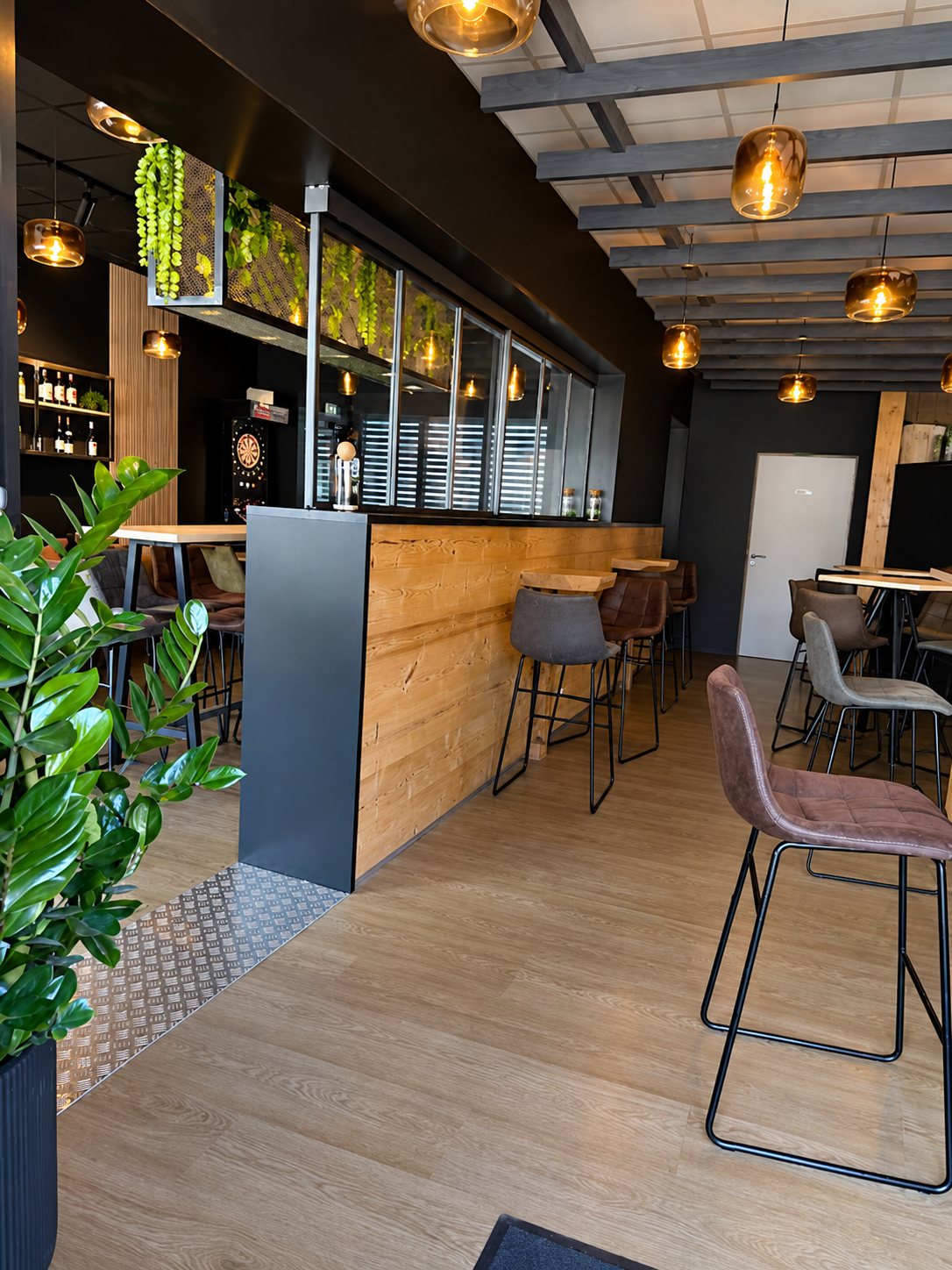 Intérieur d'un bar moderne avec un comptoir en bois et métal, des tabourets en cuir, des suspensions ambrées et une plante verte au premier plan.