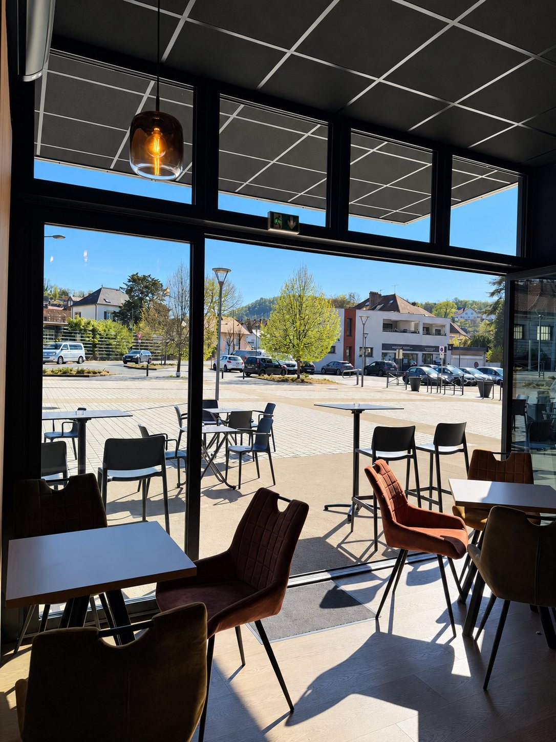 Salle de café avec tables et chaises en velours marron, grande baie vitrée ouverte sur la terrasse et la place ensoleillée.
