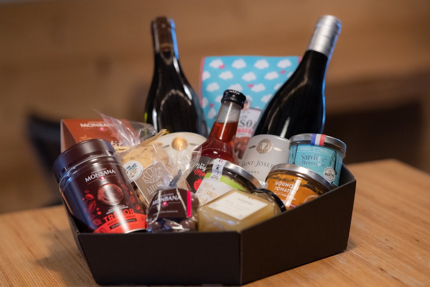 Un panier cadeau noir posé sur une table en bois contient deux bouteilles de vin, du chocolat Monbana, des rillettes de sardine et d'autres produits gourmands.