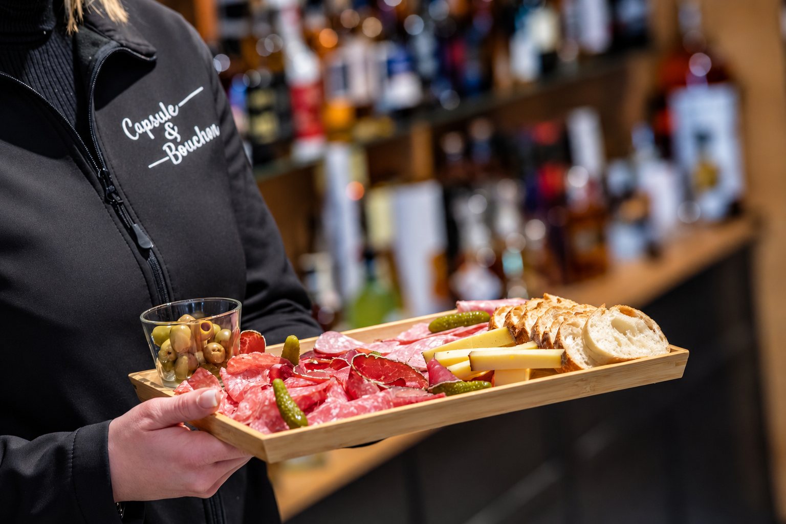 Une serveuse en veste noire brodée tient un plateau en bambou garni de charcuterie, fromage, pain, cornichons et olives devant des étagères de bouteilles floues.