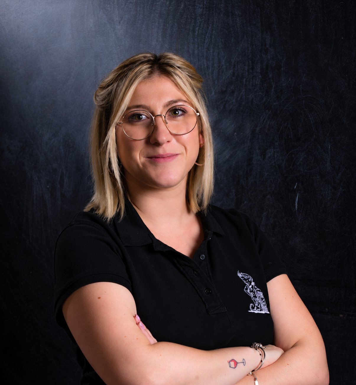 Une jeune femme blonde portant des lunettes rondes et un polo noir brodé sourit, les bras croisés, devant un fond sombre.