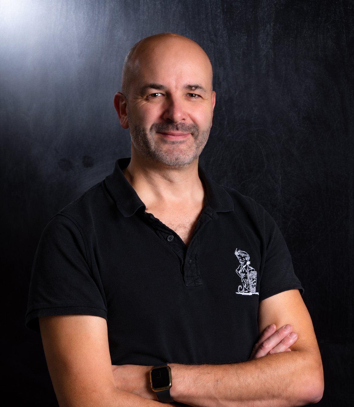 Un homme chauve et souriant, vêtu d'un polo noir brodé d'un petit personnage, pose les bras croisés devant un fond sombre.