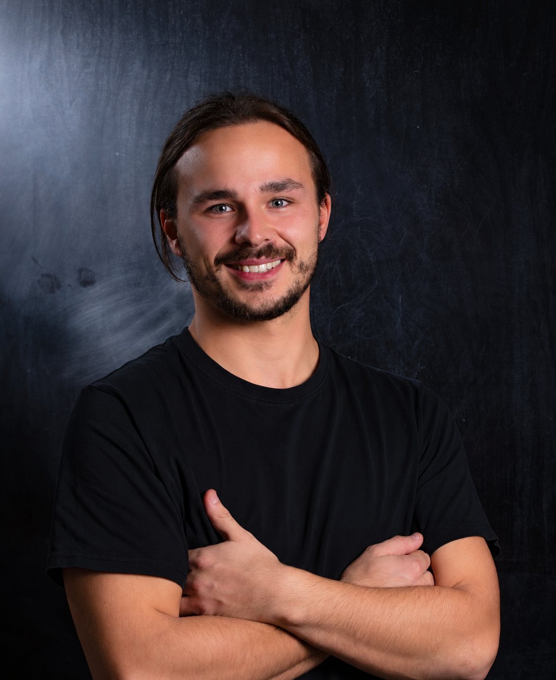 Portrait d'un jeune homme souriant, barbu, les cheveux attachés, vêtu d'un t-shirt noir et les bras croisés devant un fond sombre.