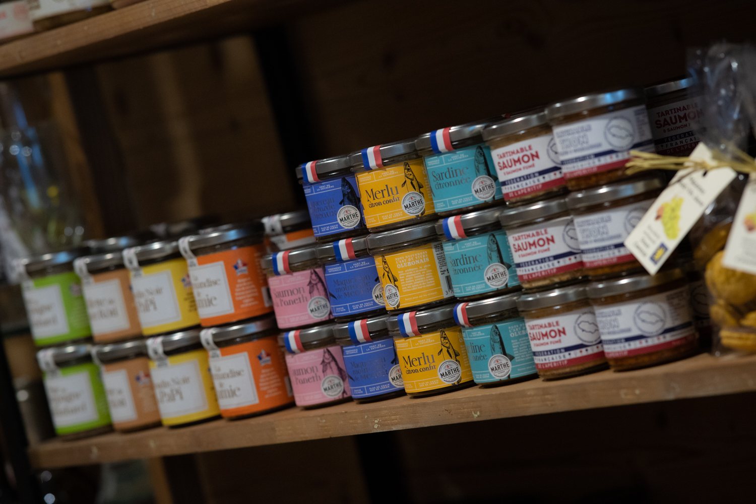 Des rangées de petits pots de tartinables de poisson, merlu, sardine et saumon, alignés sur une étagère en bois dans une épicerie.