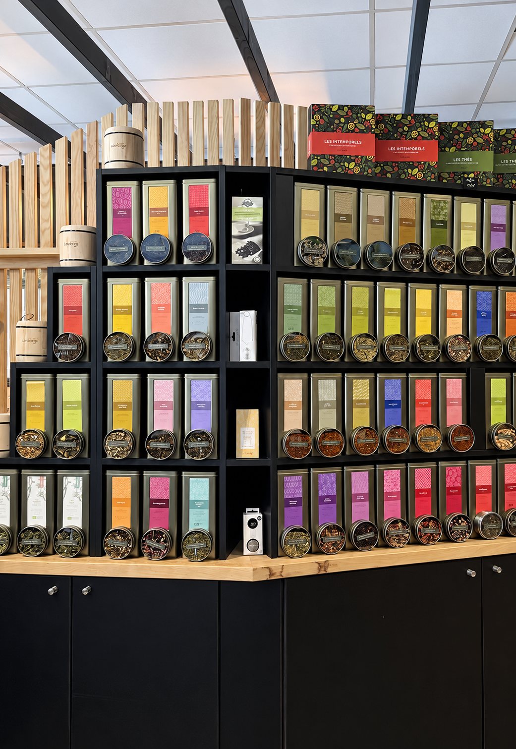 Un meuble présentoir noir avec plateau en bois expose de nombreuses boîtes de thé aux étiquettes colorées, surmontées de coffrets et d'une cloison en bois clair.
