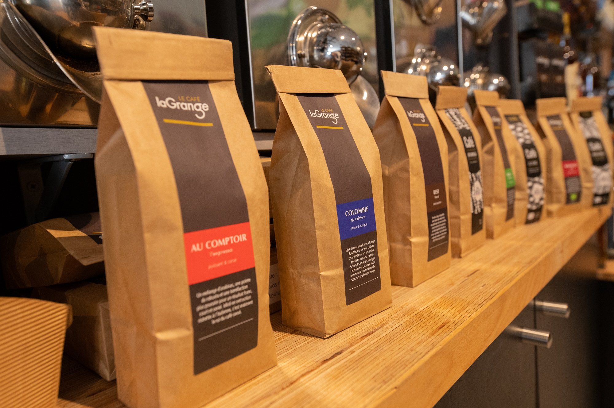 Une rangée de sachets de café en papier kraft de la marque La Grange, dont Au Comptoir et Colombie, posés sur un comptoir en bois clair.