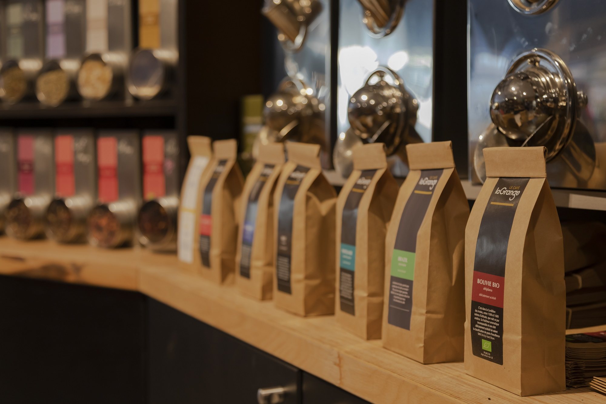 Une rangée de sachets de café en papier kraft avec étiquettes colorées posés sur un comptoir en bois, devant des distributeurs de thé en vrac.