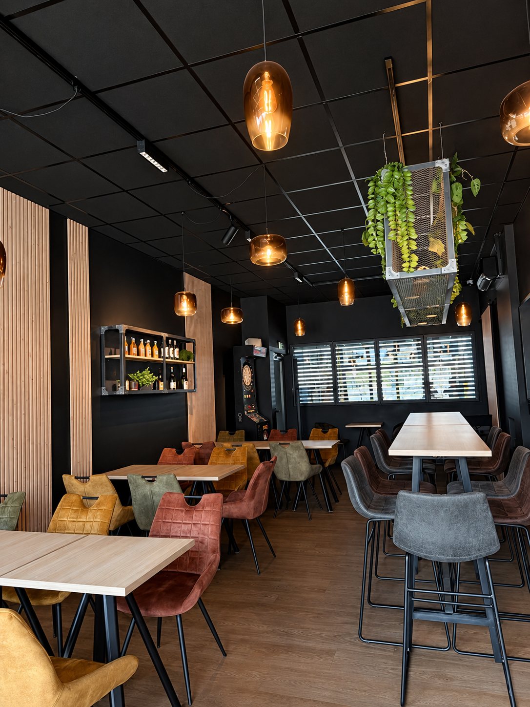 Intérieur d'un bar aux murs noirs et panneaux de bois clair, avec tables hautes, chaises en velours colorées, suspensions ambrées et plantes suspendues.
