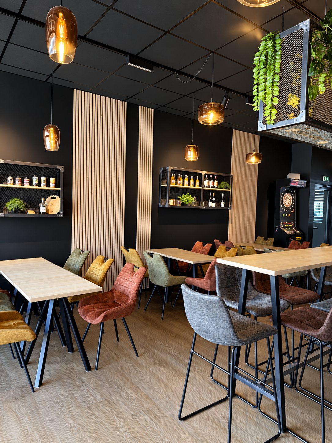 Intérieur d'un bar aux murs noirs et lattes de bois, avec tables hautes, chaises en cuir colorées, suspensions ambrées et une cible de fléchettes au fond.