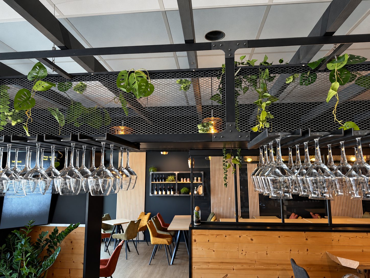 Intérieur d'un restaurant moderne avec des verres à pied suspendus au-dessus du bar, des plantes retombant d'une structure métallique et des tables en bois avec chaises colorées.