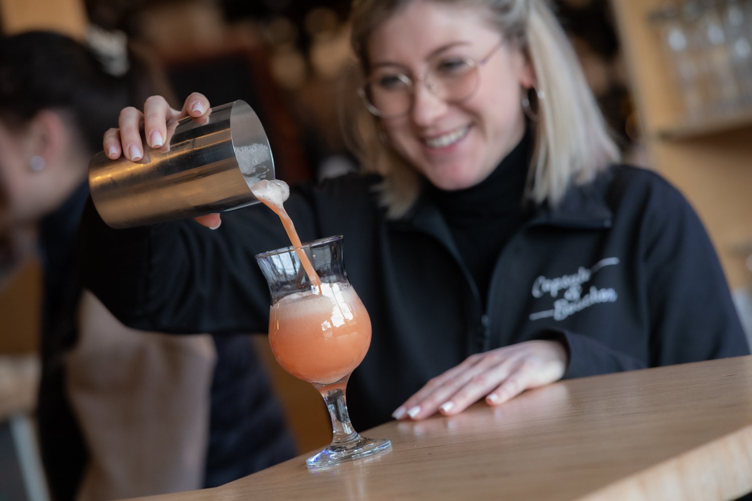 Une serveuse souriante à lunettes verse un cocktail rose mousseux depuis un shaker dans un verre à pied posé sur un comptoir en bois.