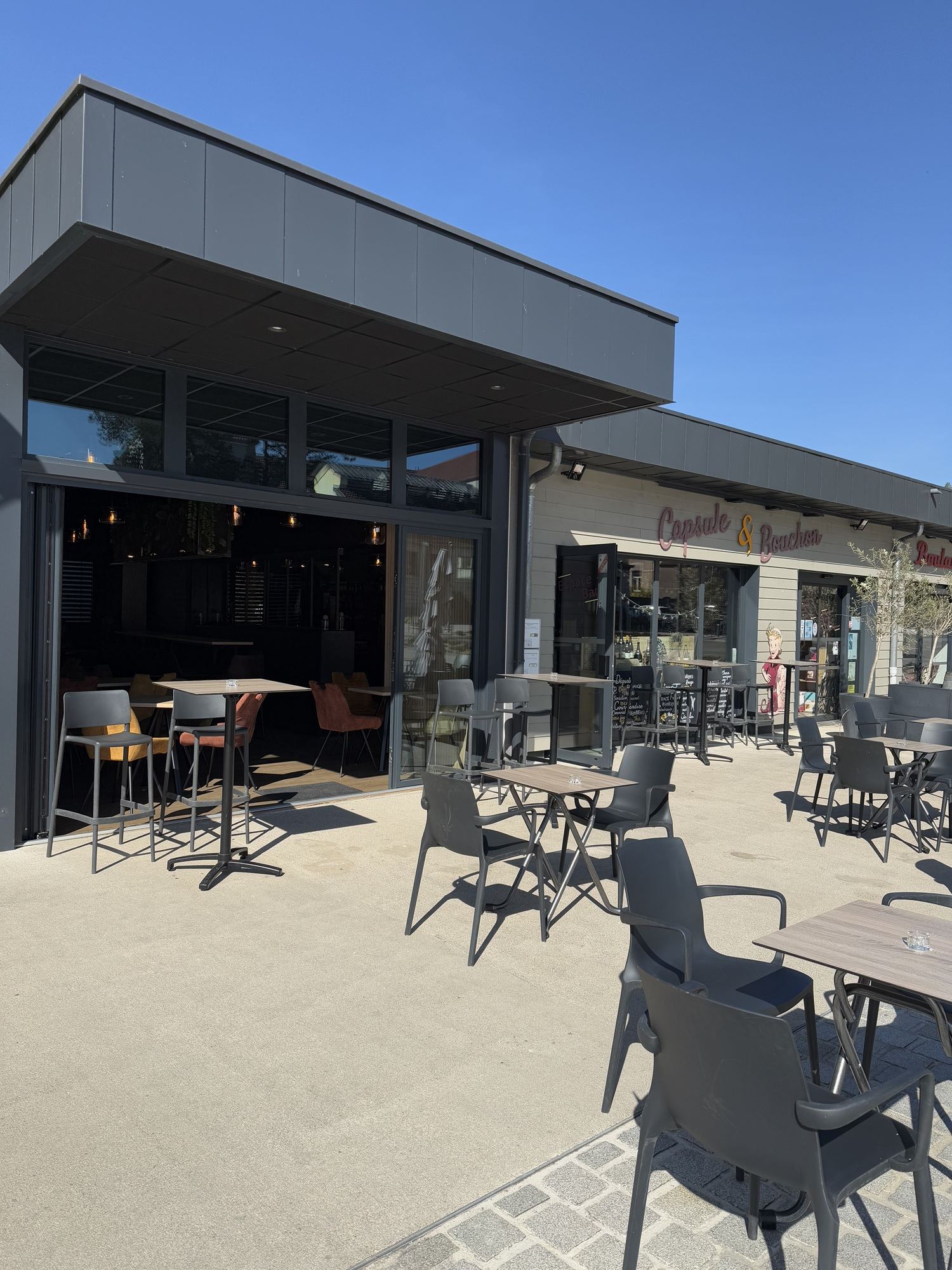 Terrasse en plein soleil devant un café-restaurant moderne, avec des tables hautes et basses et des chaises grises, et une enseigne rose sur la façade.
