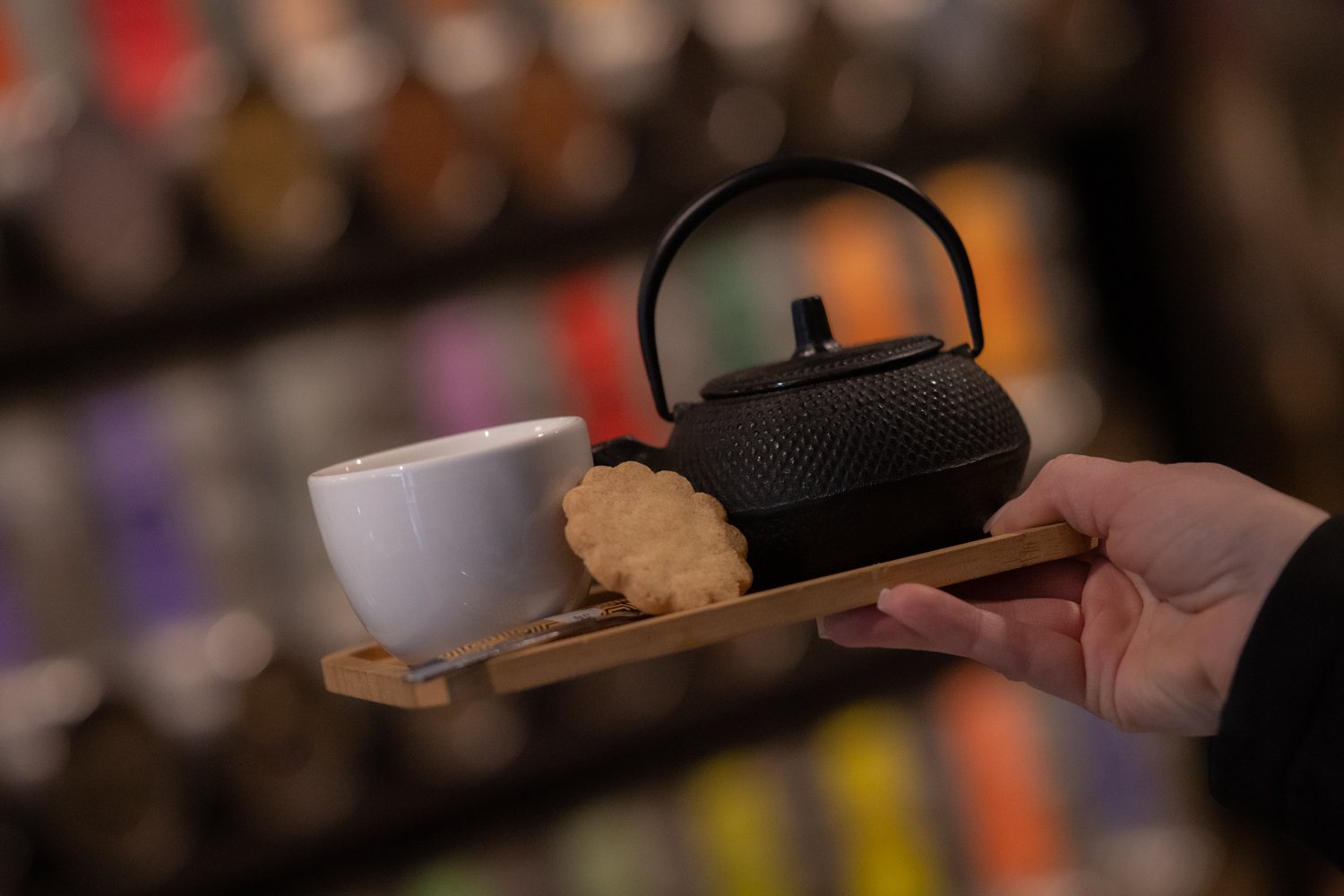 Une main tient un petit plateau en bambou portant une théière en fonte noire, une tasse blanche et un biscuit sablé, devant des rangées de boîtes de thé floues.