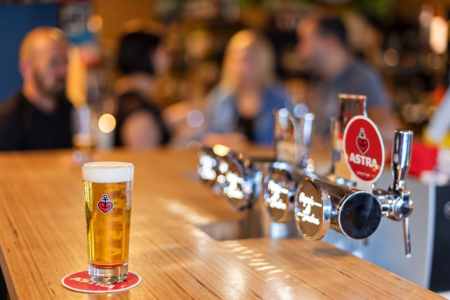 Un verre de bière blonde Astra posé sur un sous-bock au bout d'un comptoir en bois, à côté de tireuses chromées, avec des clients flous à l'arrière-plan.
