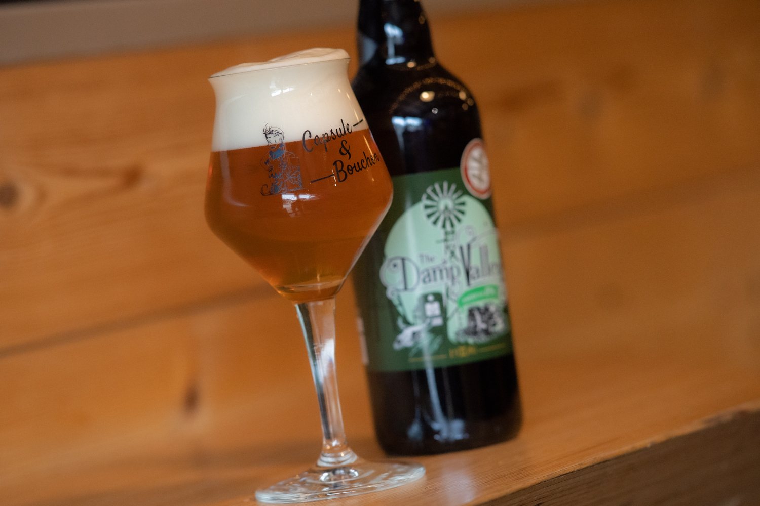 Un verre à pied rempli de bière blonde avec une belle mousse, posé à côté d'une bouteille à étiquette verte sur une surface en bois.
