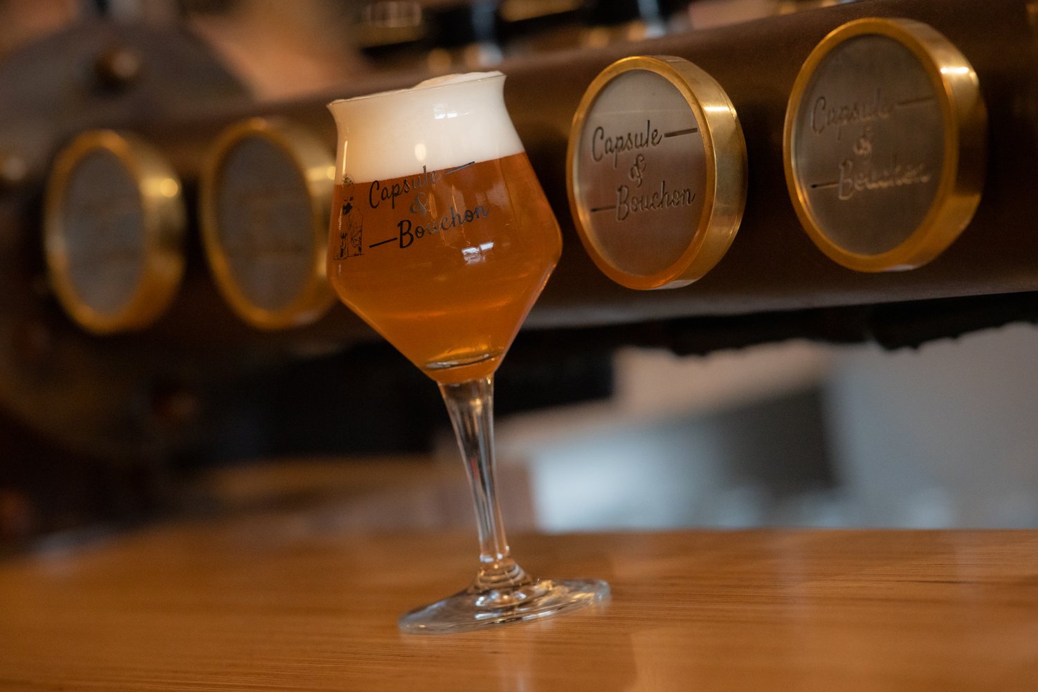 Un verre à pied rempli de bière blonde avec une belle mousse, posé sur un comptoir en bois devant des médaillons en laiton gravés Capsule & Bouchon.